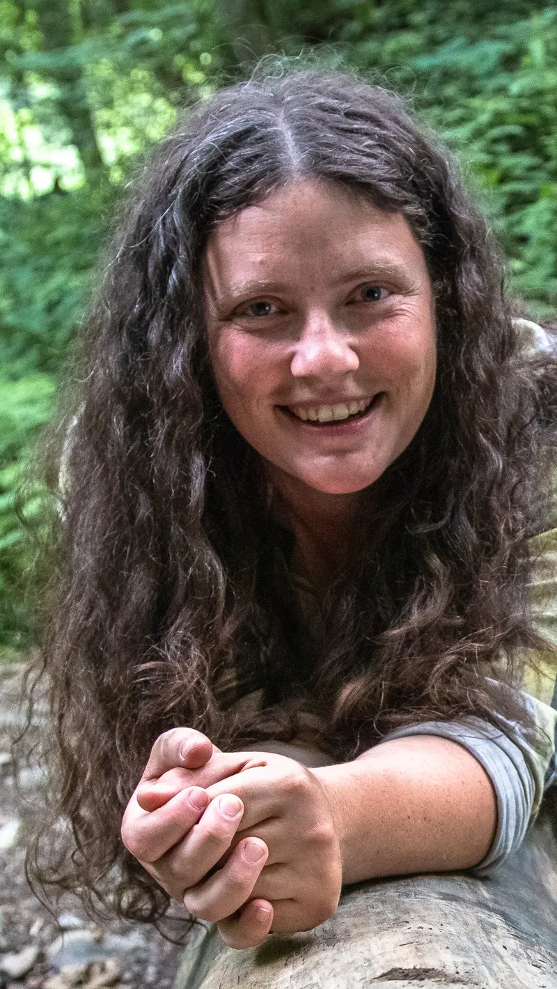 Anna Skryjová opřená o padlý kmen v lese, ruce sepjaté, usmívá se